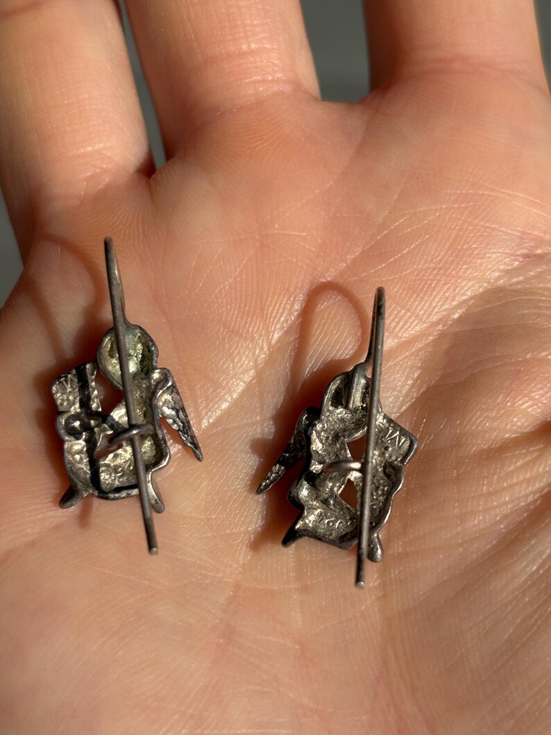 May include: A pair of silver earrings with a detailed design of a seated winged figure, possibly an angel or cherub. The earrings have a post and hook closure, and the design includes intricate detailing.