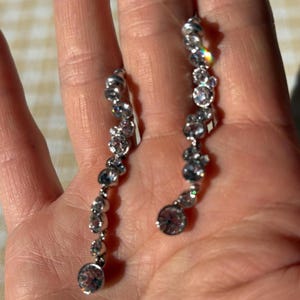 May include: A pair of silver-toned dangle earrings. Each earring features a series of clear, round, and faceted gemstones of varying sizes, arranged in a vertical line. The gemstones catch the light, creating a sparkling effect. The earrings are displayed against a skin-toned background.