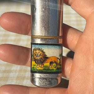 May include: A vintage silver metal match safe with a rectangular image of a lion. The match safe has a cylindrical shape with a hinged lid and a gold-colored band. The lion is depicted in shades of orange, yellow, and brown.