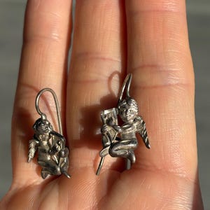 May include: A pair of silver-coloured earrings, each depicting a cherub. One cherub is seated and playing a harp, the other also seated, holding a harp. The earrings have a hook closure.