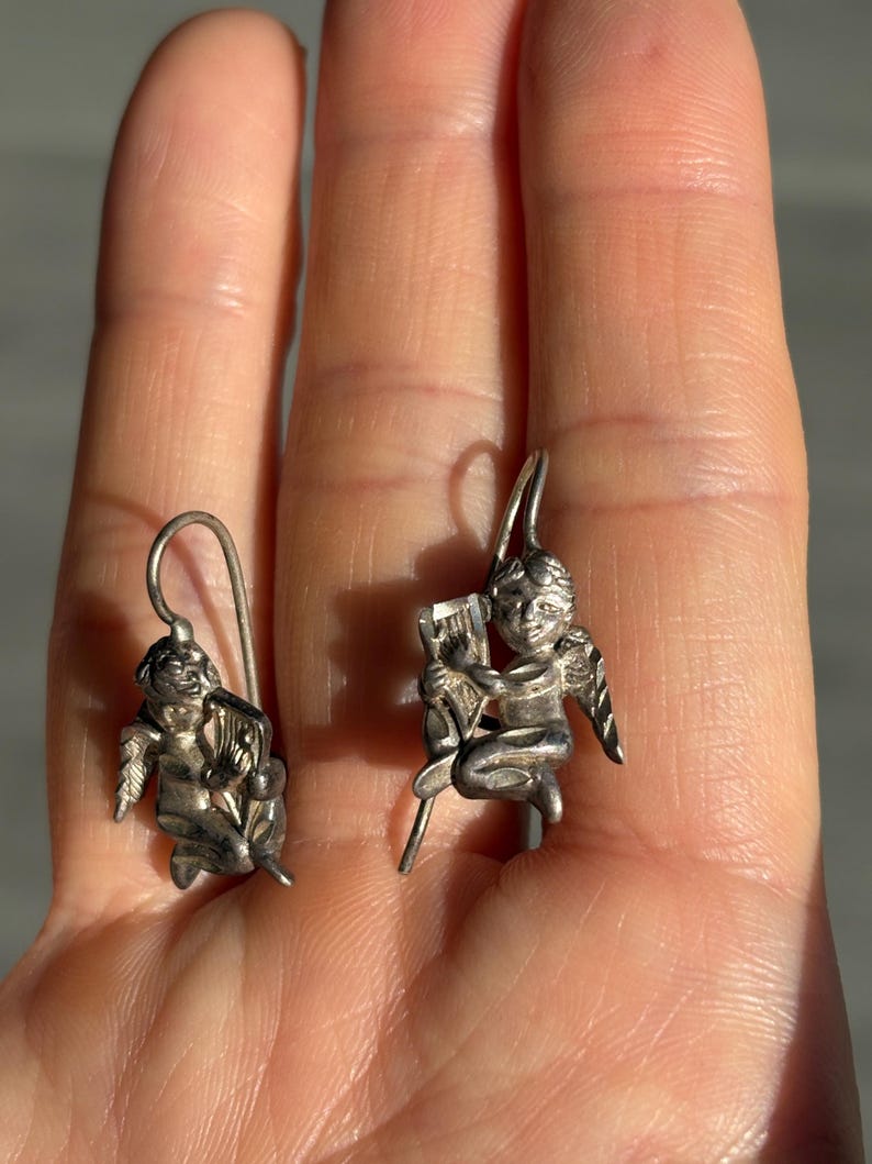 May include: A pair of silver-coloured earrings, each depicting a cherub. One cherub is seated and playing a harp, the other also seated, holding a harp. The earrings have a hook closure.
