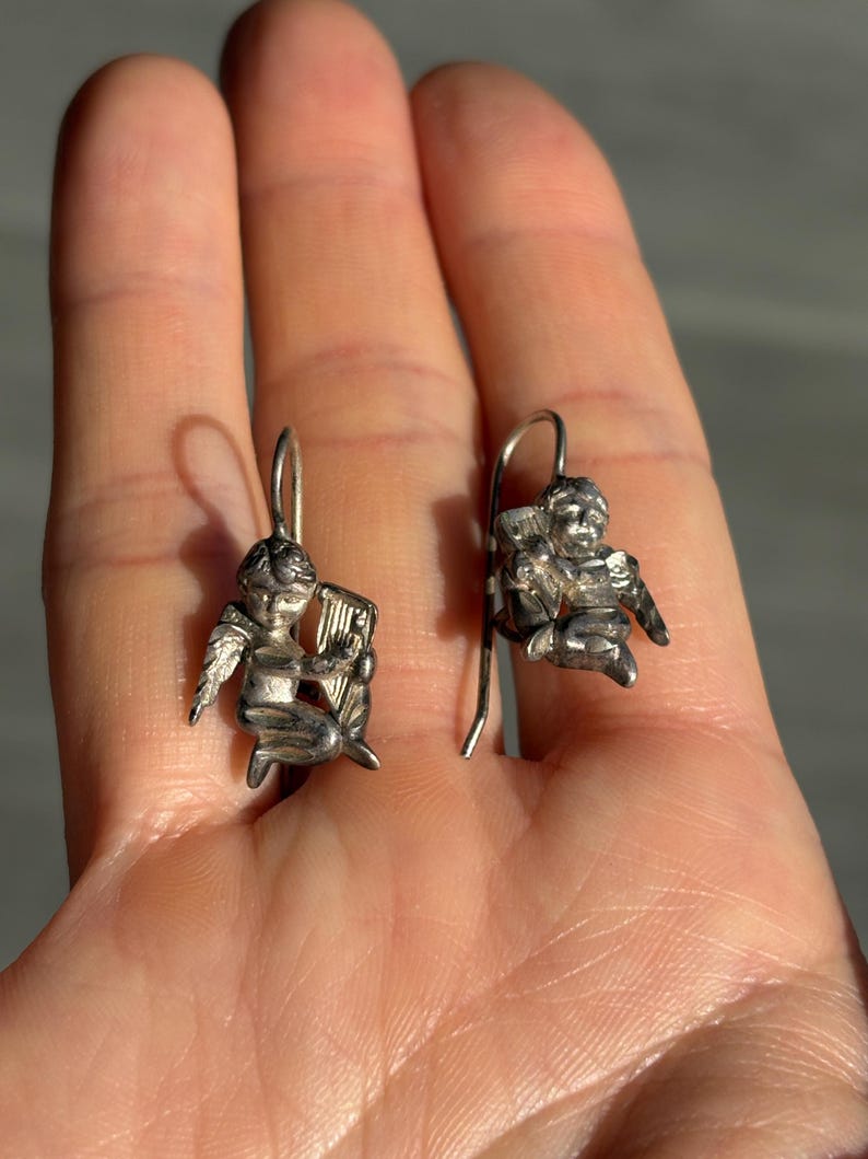 May include: Two silver-coloured earrings, each depicting a small cherub playing a harp. The cherubs have detailed wings and are suspended from simple ear wires. The earrings are held in a hand, showing their size and design.