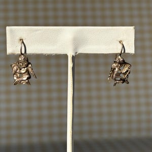 May include: A pair of silver-coloured angel earrings. Each earring features a detailed cherub design with outstretched wings and a seated pose. The earrings are displayed on a white earring stand against a neutral background.