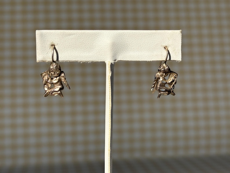 May include: A pair of silver-coloured angel earrings. Each earring features a detailed cherub design with outstretched wings and a seated pose. The earrings are displayed on a white earring stand against a neutral background.