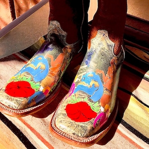 May include: A pair of cowboy boots with a colorful, artistic design. The boots have a dark brown shaft and a painted footbed featuring red, blue, and orange abstract shapes. The boots are displayed on a striped textile.