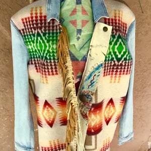 May include: A unique jacket with a denim collar and sleeves, featuring a colorful geometric pattern in red, green, and orange on a cream background. The jacket has a fringed leather accent and a patterned lining.