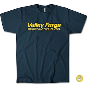 May include: A navy blue t-shirt featuring the yellow text "Valley Forge AUTOMOTIVE CENTER". The text is centrally placed. The shirt has a crew neck and short sleeves.