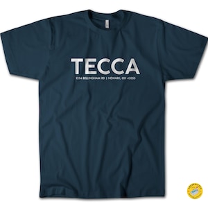 Puede incluir: Camiseta azul marino con la palabra "TECCA" en letras blancas. Debajo, la dirección "1334 Bellingham Rd | Newark, OH 43055" también en blanco. La camiseta tiene mangas cortas y cuello redondo.