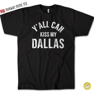 May include: A black short-sleeved t-shirt featuring white text printed on the chest that reads Y'ALL CAN KISS MY DALLAS in a bold, sans-serif font. The shirt has a crew neckline and a standard fit, with the text centered across the front.