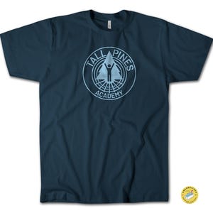 Peut inclure: T-shirt bleu marine avec un motif bleu clair. Le motif présente le texte "TALL PINES ACADEMY" dans un design circulaire, avec un arbre stylisé et une figure au centre. Le t-shirt est en matière souple.