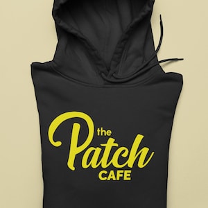May include: Black hooded sweatshirt with the text "the Patch CAFE" in yellow script. The hoodie has a drawstring and a hood. The sweatshirt is folded on a light beige surface.