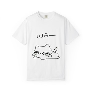 Könnte beinhalten: Weißes T-Shirt mit einer schwarzen Cartoon-Katzen-Illustration und dem Wort "WA-". Die Katze ist in einem einfachen Stil mit geschlossenen Augen gezeichnet, was eine entspannte Stimmung vermittelt. Das T-Shirt hat einen klassischen Rundhalsausschnitt.