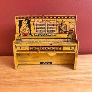 May include: A vintage yellow toy piano with black keys and decorative illustrations. The piano features the text "THE OGAATCH" and musical notes. A pig illustration is on the left side, and a framed picture and lamp are on the right.