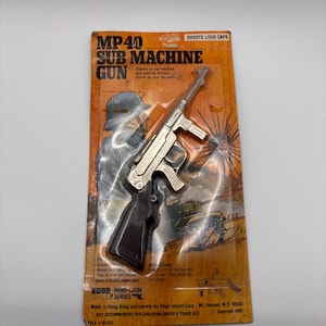 May include: A vintage toy MP40 submachine gun replica on its original packaging. The packaging is orange and brown with illustrations of soldiers and tanks. The toy gun is silver and black, and the packaging reads "SHOOTS LOUD CAPS."