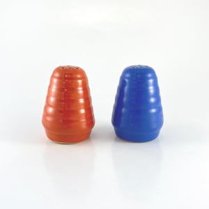 May include: A set of two ceramic salt and pepper shakers. The shaker on the left is a vibrant orange color, while the shaker on the right is a bright blue color. Both shakers have a ridged, conical shape.