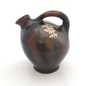 May include: A small brown ceramic pitcher with a floral design. The pitcher has a handle and a spout.