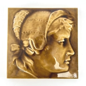 May include: A square, brown ceramic tile featuring a bas-relief profile of a person wearing a bonnet. The tile has a glossy finish and is a warm, earthy brown color. The person's profile is detailed, with visible facial features and the texture of the bonnet.