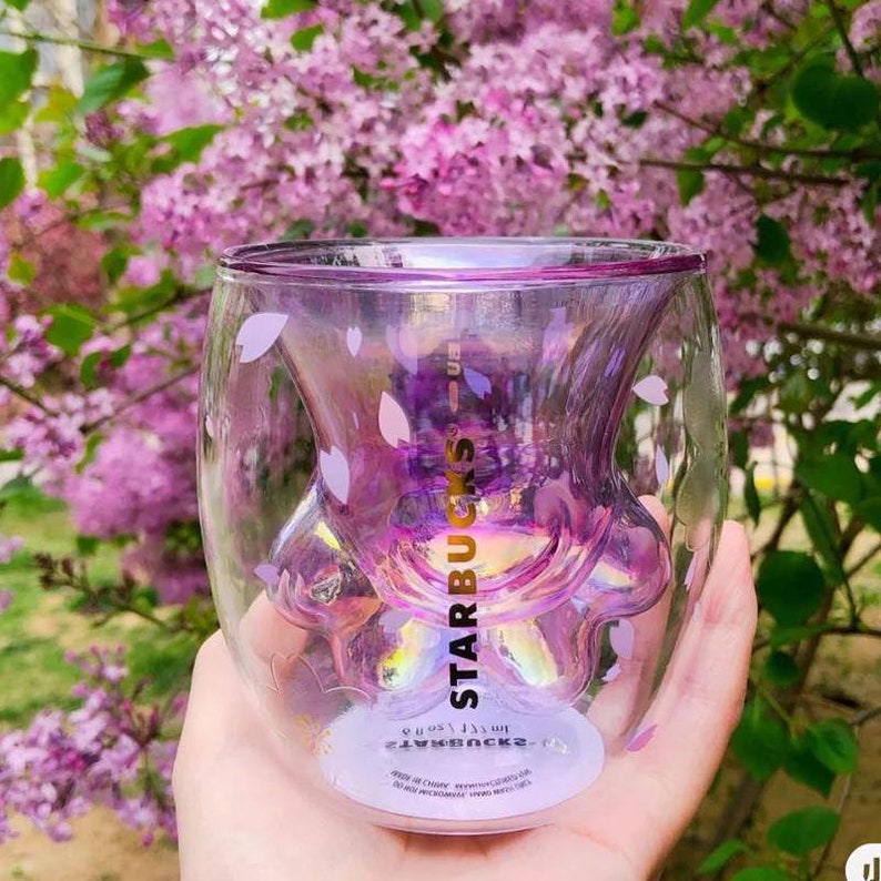 May include: A clear glass double-walled mug with a paw print design. The mug is iridescent and has a pink tint. The mug has the Starbucks logo and the text "STARBUCKS" on the side. The mug is decorated with small pink flower designs.