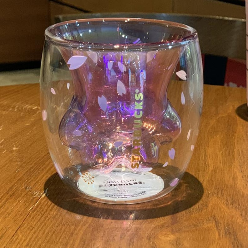 May include: A double-walled glass mug with a paw print design and pink cherry blossom accents. The mug features the Starbucks logo and the text "Enjoy your Starbucks."