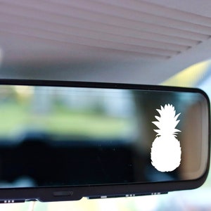May include: A white pineapple decal is affixed to a car's rearview mirror. The pineapple is a silhouette, with a detailed crown and textured body. The mirror reflects a blurred outdoor scene, suggesting a sunny day.