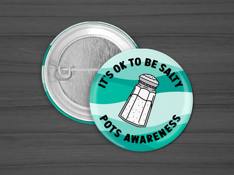 Puede incluir: Un bot&oacute;n azul verdoso y blanco con el texto "It's OK to be Salty" y un gr&aacute;fico de un salero. El texto "POTS Awareness" tambi&eacute;n est&aacute; en el bot&oacute;n.