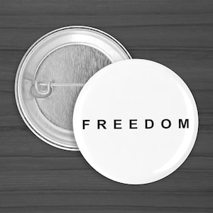 May include: A white button with the word "FREEDOM" in black, with a silver metal backing. The button is round and has a pin on the back. The button is on a dark gray wooden surface.