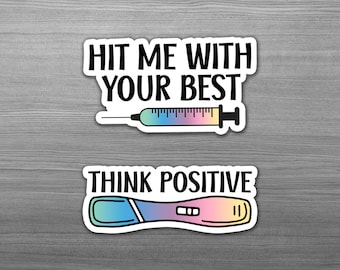 May include: Two stickers with a humorous theme. The top sticker features a syringe with a rainbow-colored needle and the text "Hit me with your best". The bottom sticker features a pregnancy test with a rainbow-colored design and the text "Think Positive".