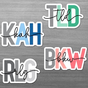 May include: Four colourful stickers with the words "KAH", "RLG", "TLd", and "BKw" written in black cursive script. The letters are outlined in white and each sticker has a different colour scheme.