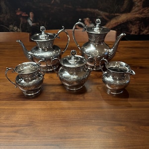 May include: A silver tea set with intricate floral patterns. The set comprises two teapots, a sugar bowl, a creamer, and a small jug. The items are displayed on a wooden surface, reflecting the light.