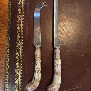 May include: Two cheese knives with ornate handles. One knife has a curved blade with a forked tip, and the other has a long, serrated blade. The handles are white with a floral design in pink, gold, and green.