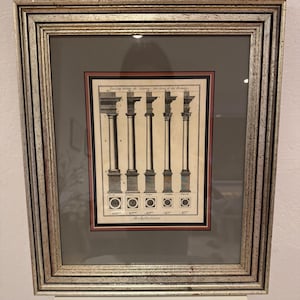 May include: Framed architectural print featuring five classical columns with detailed drawings of their capitals and bases. The print is in black and white with a silver-toned frame and a gray mat. The word "Architecture" is printed at the bottom.