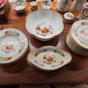 May include: A collection of antique porcelain tableware with floral designs. Includes plates, bowls, and serving dishes with orange, blue, and gold accents. The set is arranged on a dark wooden surface.