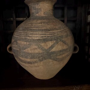 May include: A large, antique ceramic vase with a wide body and a flared rim. The vase is tan with dark gray geometric patterns and horizontal stripes. It has two small handles and is likely an ancient artifact.