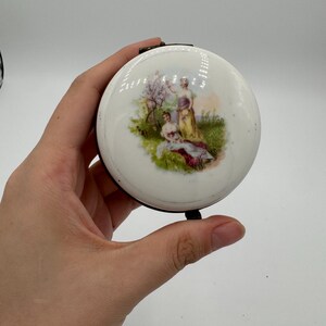 Puede incluir: Un estuche compacto redondo blanco con un marco de metal negro. La tapa presenta una escena pintada de tres figuras en un jardín con un árbol y hierba. La obra de arte tiene una estética vintage.