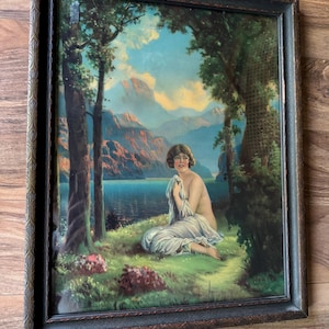 May include: Framed vintage print depicting a woman seated near a lake. The artwork features a serene landscape with mountains, trees, and a body of water. The frame is dark brown with an ornate design. The woman is draped in white fabric.