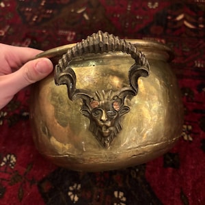 Puede incluir: Una olla redonda de latón con un asa prominente en forma de cabeza de león estilizada con detalles ornamentados y curvos. La superficie metálica tiene una pátina cálida de color marrón dorado con signos visibles de antigüedad y desgaste.