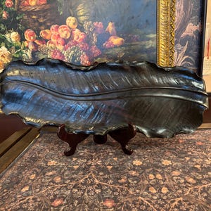 May include: A long, dark brown decorative tray shaped like a large, elongated leaf with a prominent central vein and rippled, organic edges. The tray is crafted with a smooth, polished finish and is displayed resting on a dark brown stand.