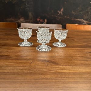 May include: Five clear glass shot glasses with a cut glass design. The glasses are arranged in a row on a wooden table.