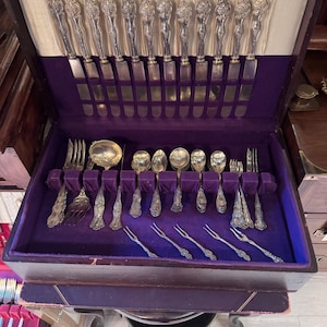 Puede incluir: Un juego de cubiertos de tono plateado guardado en un cofre de madera oscura con interior de terciopelo morado. El juego incluye cuchillos con mangos ornamentados, varias cucharas, tenedores y pequeños tenedores para mariscos.