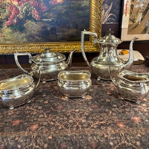 May include: A silver tea service set consisting of a teapot, a coffee pot, a sugar bowl, and two creamers. Each piece features a polished metallic finish, a rounded body, and a beaded rim detail. The set is arranged on a dark floral patterned surface.