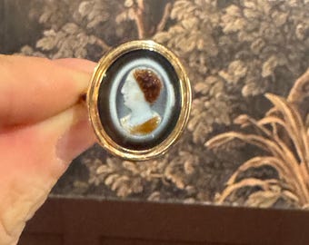 Puede incluir: Un anillo dorado con un retrato de camafeo de una mujer con cabello castaño y rostro blanco. El camafeo está engastado sobre un fondo negro dentro de un marco circular. El anillo se sostiene contra un fondo estampado.