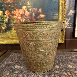 May include: A tapered brass bucket featuring intricate embossed patterns of horses and foliage. The metal surface displays a weathered patina with decorative bands of repeating leaf motifs along the top and bottom edges and a single metal handle.
