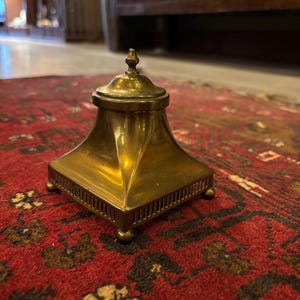 May include: A brass inkwell with a square base and a pyramidal body. The inkwell has a lid with a small finial. The piece is made of polished brass and sits on a red rug with a dark pattern. The inkwell is an antique.