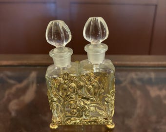 May include: A vintage perfume bottle with two clear glass containers and stoppers. The bottles are encased in a gold-colored filigree with floral designs. The perfume bottle sits on a reflective surface.
