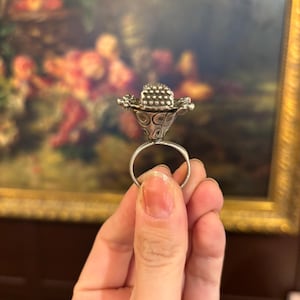 Op de afbeelding: Een zilveren ring met een driedimensionale bloemvormige bovenkant. Het midden van de bloem heeft een getextureerd, gestippeld oppervlak, terwijl de basis van de bloemkop een reeks gegraveerde cirkelvormige patronen vertoont op de ring.