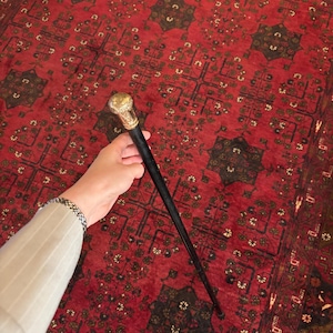 Op de afbeelding: Een wandelstok met een lange, slanke zwarte schacht en een afgerond, goudkleurig metalen handvat met ingewikkelde reliëfdetails. Het handvat heeft een gepolijste afwerking, terwijl de schacht glad is en taps toeloopt naar een kleine, donkere punt.