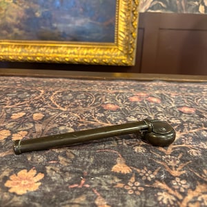 May include: A vintage, dark bronze-colored pen with a rounded end and a small, circular cap. The pen is lying on a patterned fabric with floral and leaf designs. The background includes a framed painting.