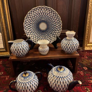 May include: A porcelain tea set featuring a blue and gold lattice pattern on a white background. The set includes a teapot, a lidded sugar bowl, a creamer, a small vase, a larger vase, and a decorative plate, all with gold-trimmed edges.