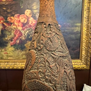 May include: A brown, textured bottle with intricate carved designs. The bottle has a narrow neck and a wider base, with a rough, earthy appearance. The detailed carvings cover the entire surface, creating a unique decorative piece.