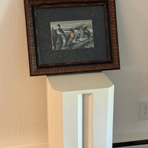 Puede incluir: Una impresión enmarcada que presenta una ilustración detallada de varios pájaros posados en ramas. La obra de arte está sobre un fondo oscuro y enmarcada en un marco de madera marrón oscuro y ornamentado. La impresión se exhibe sobre un pedestal blanco.